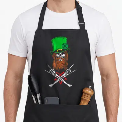 Tablier de cuisine noir avec crâne barbu, chapeau vert et os croisés en motif central.