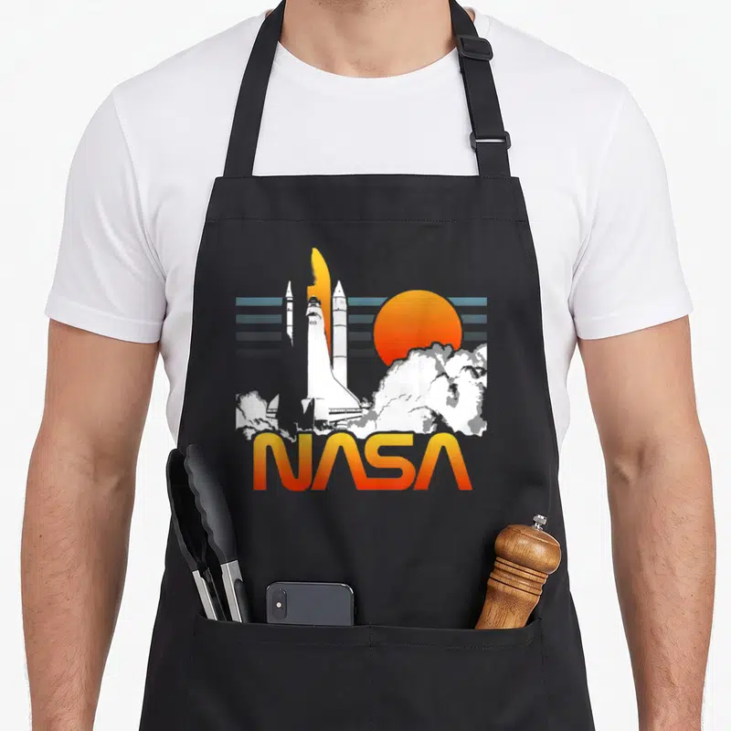 Tablier de cuisine noir avec illustration de fusée spatiale, nuages de fumée et logo NASA en lettres orange et jaunes.