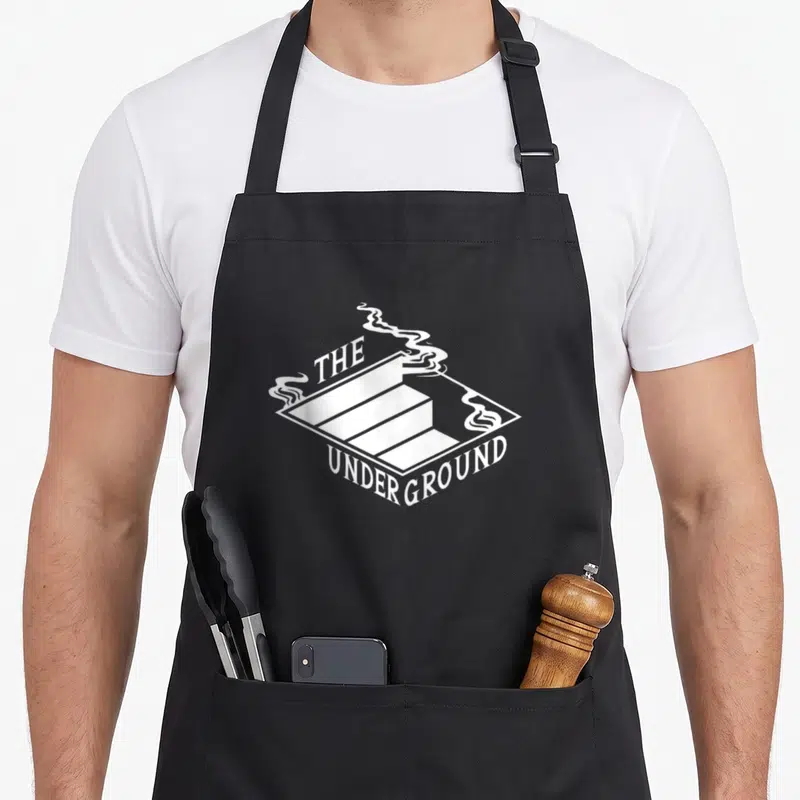 Tablier de cuisine noir avec logo blanc The Underground et fumée s'élevant d'une structure géométrique en perspective.