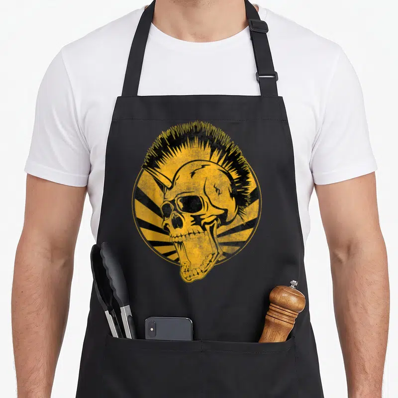 Tablier de cuisine noir avec un crâne punk au mohawk jaune et des rayons noirs en arrière-plan circulaire.