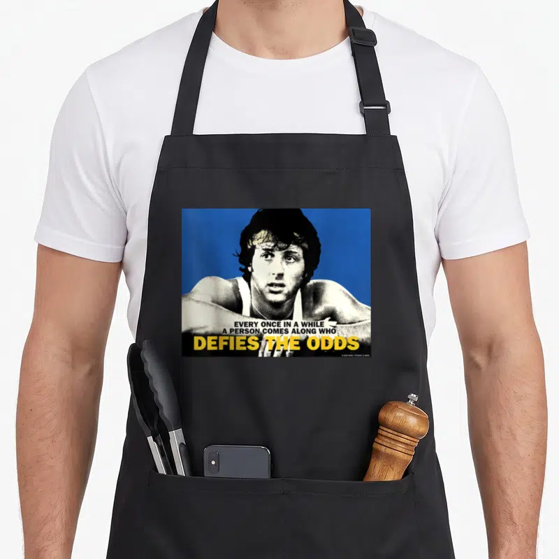 Tablier de cuisine noir avec une illustration de Rocky sur fond bleu et texte jaune Defies the Odds.