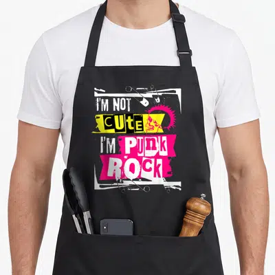 Tablier de cuisine noir avec inscription « I'm Not Adouci I'm Punk Rock », crâne rose et épingles de sécurité sur fond blanc et jaune.