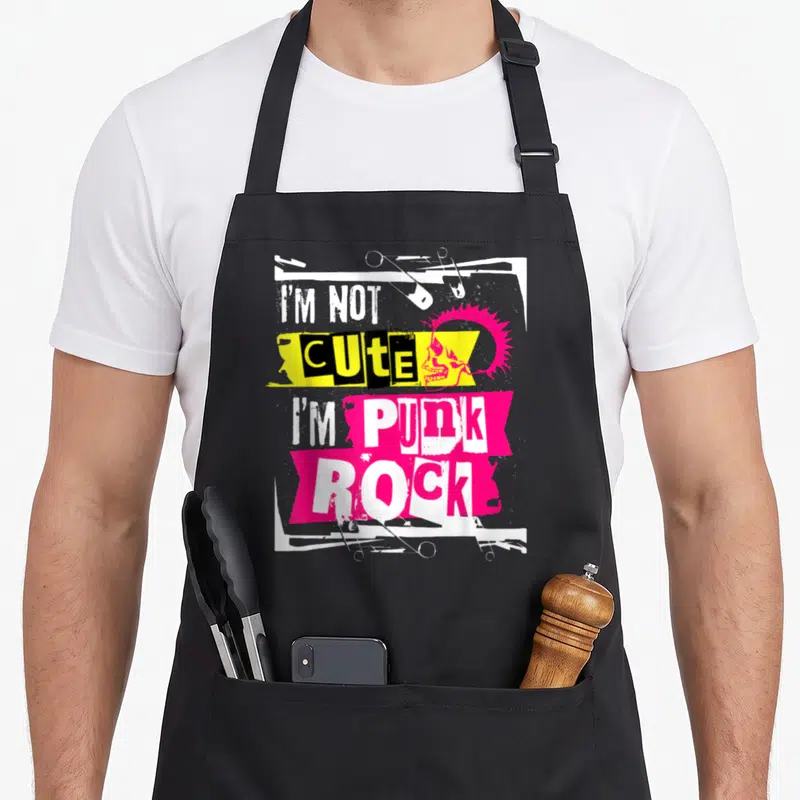 Tablier de cuisine noir avec inscription « I'm Not Adouci I'm Punk Rock », crâne rose et épingles de sécurité sur fond blanc et jaune.