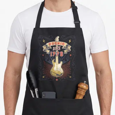 Tablier de cuisine noir avec illustration vintage guitare électrique ailes et texte Rockin Since 1978 encadré de volutes décoratives.