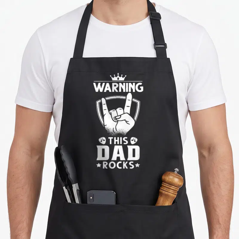 Tablier de cuisine noir avec l'inscription blanche Warning This Dad Rocks et un geste de la main heavy metal.