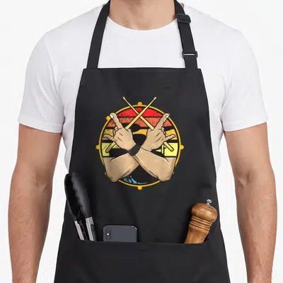 Tablier de cuisine noir avec illustration vintage de baguettes de batterie croisées et gestes de rock sur fond de drapeau aux couleurs vives.