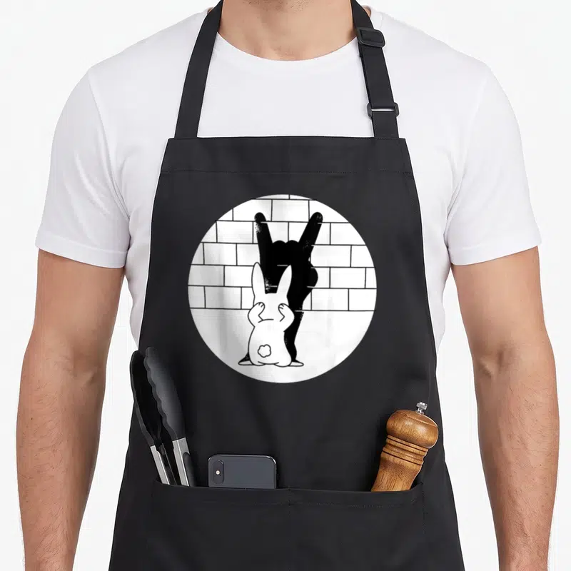 Tablier de cuisine noir avec illustration d'un lapin blanc face à une main noire formant le signe du metal contre un mur de briques.