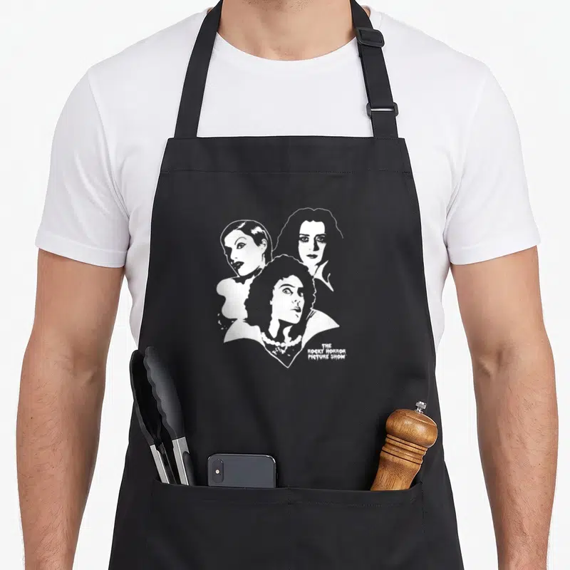 Tablier de cuisine noir avec illustration blanche représentant le trio du Rocky Horror Picture Show avec Col en V imprimé en bas à droite.
