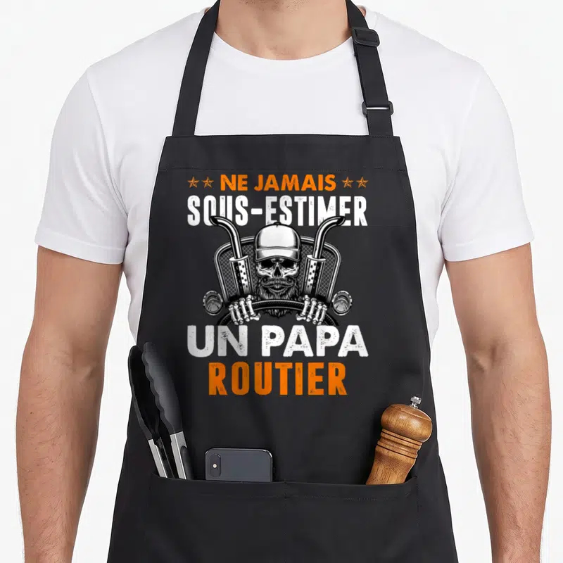 Tablier de cuisine noir avec un crâne barbu coiffé d'une casquette entre deux tuyaux d'échappement et le texte Ne jamais sous-estimer un papa routier.