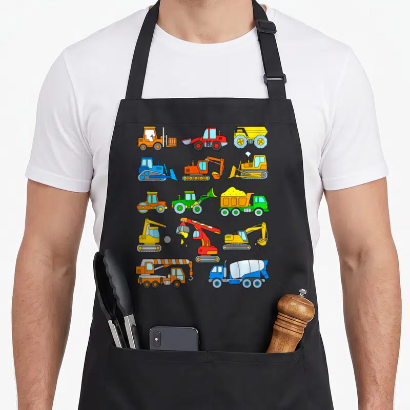 Tablier de cuisine noir avec illustrations colorées de camions, pelles mécaniques et engins de chantier disposés en grille.