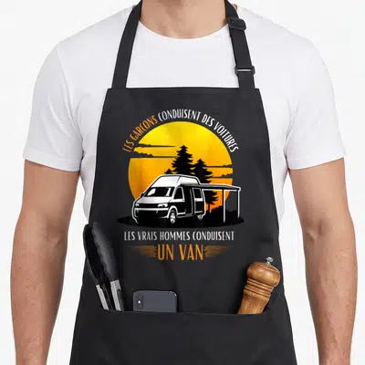 Tablier de cuisine noir avec illustration d'un van blanc et texte humoristique sur fond de soleil orange.
