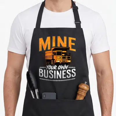 Tablier de cuisine noir avec illustration de camion de mine orange et texte MINE YOUR OWN BUSINESS en typographie robuste.