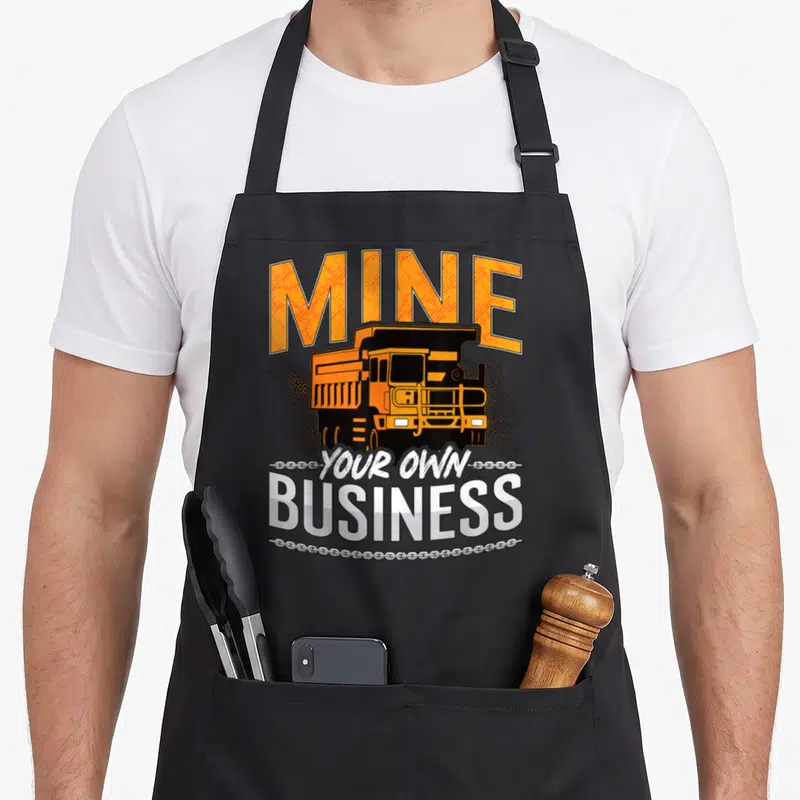 Tablier de cuisine noir avec illustration de camion de mine orange et texte MINE YOUR OWN BUSINESS en typographie robuste.