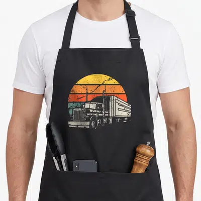 Tablier de cuisine noir avec illustration rétro d'un gros camion blanc sur fond de soleil couchant multicolore.
