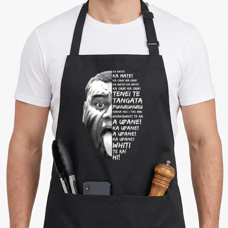 Tablier de cuisine noir avec portrait monochrome d'un joueur effectuant le Haka et des textes en maori alignés à droite.