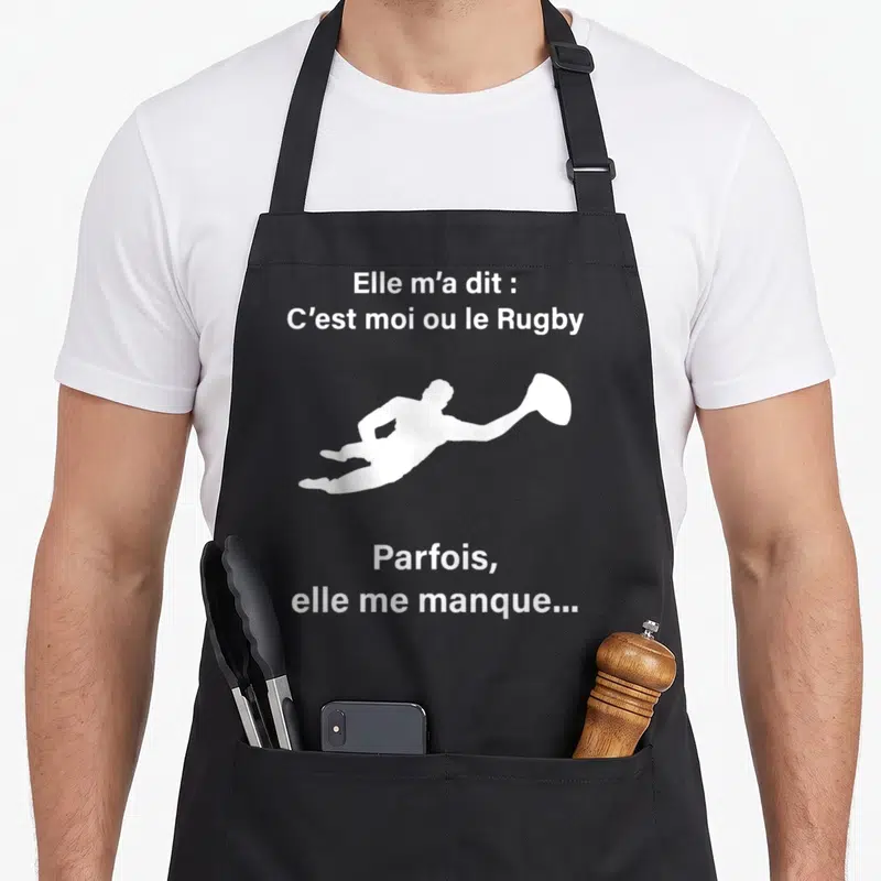 Tablier de cuisine noir avec silhouette blanche d'un joueur de rugby et texte humoristique sur fond uni.