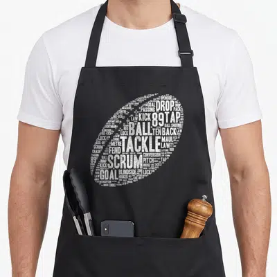 Tablier de cuisine noir avec un ballon de rugby stylisé composé de mots blancs liés au rugby.