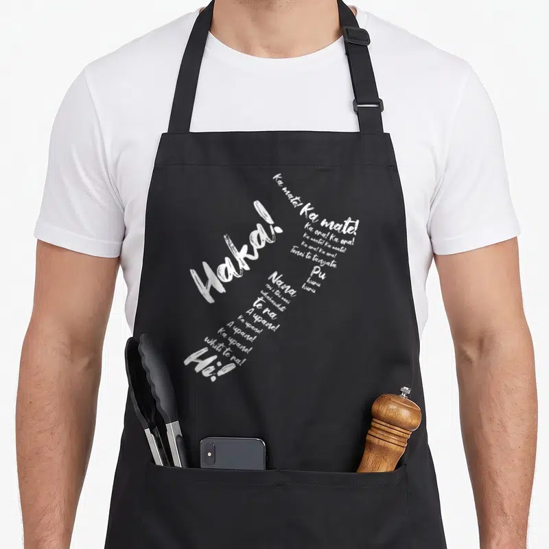 Tablier de cuisine noir avec le mot Haka et des phrases en langue Maori imprimées en blanc.