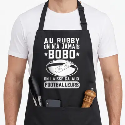 Tablier de cuisine noir avec texte blanc et illustration de ballon de rugby portant un pansement au centre.