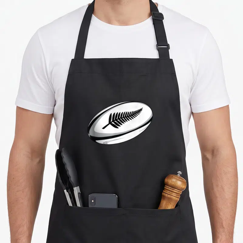 Tablier de cuisine noir avec illustration d'un ballon de rugby blanc portant une fougère noire stylisée au centre.