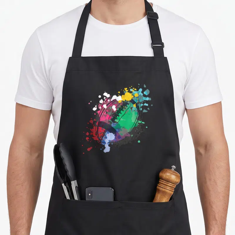 Tablier de cuisine noir avec illustration artistique d'un ballon de rugby aux éclats de peinture colorés.