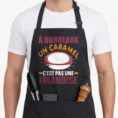 Tablier de cuisine noir avec un ballon de rugby rouge blanc et bleu et le texte À Bordeaux un caramel c'est pas une friandise.