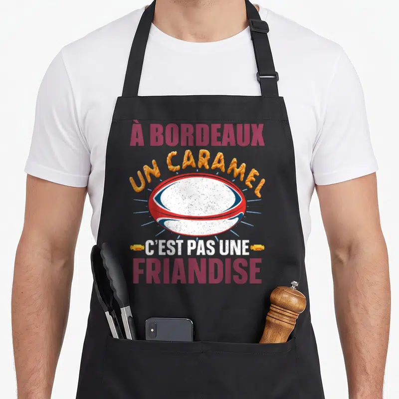 Tablier de cuisine noir avec un ballon de rugby rouge blanc et bleu et le texte À Bordeaux un caramel c'est pas une friandise.