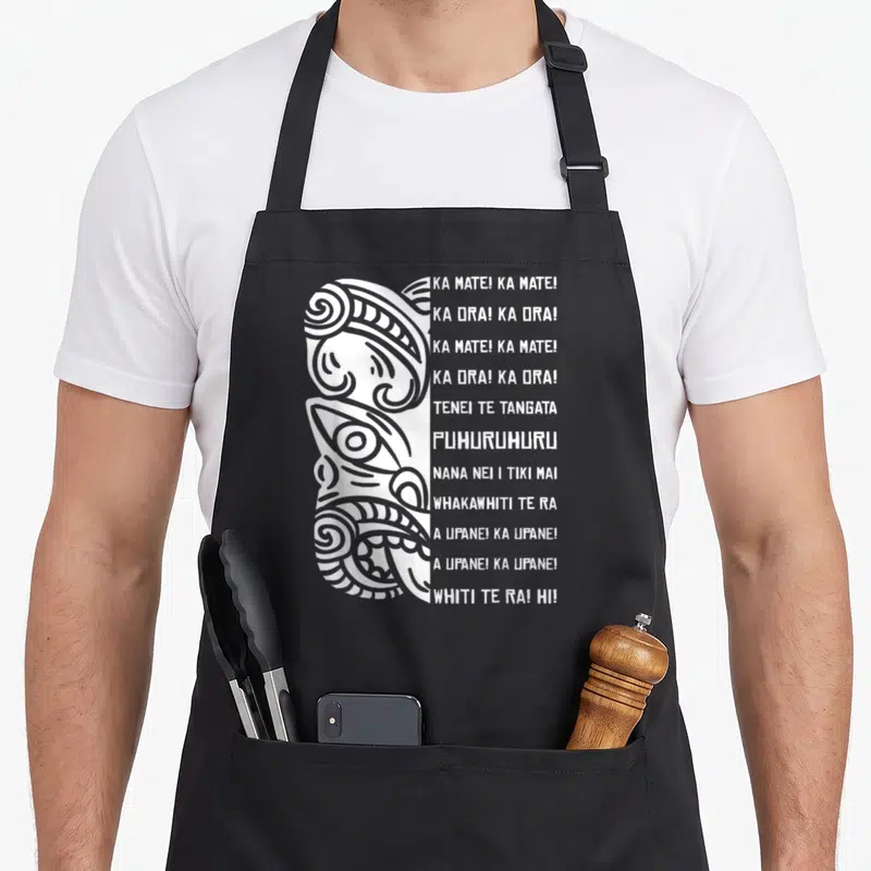Tablier de cuisine noir avec illustration blanche de visage Maori et paroles Haka alignées verticalement à droite.