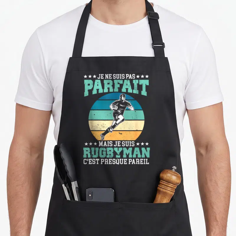 Tablier de cuisine noir avec un joueur de rugby courant sur fond circulaire coloré et texte humoristique en typographie vintage.
