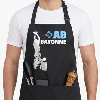 Tablier de cuisine noir avec silhouette blanche d'un joueur de rugby levant la balle et le logo AB Bayonne en bleu et blanc.