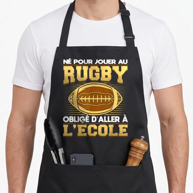 Tablier de cuisine noir avec un ballon de rugby doré et le texte né pour jouer au rugby obligé d'aller à l'école imprimé en blanc et or.