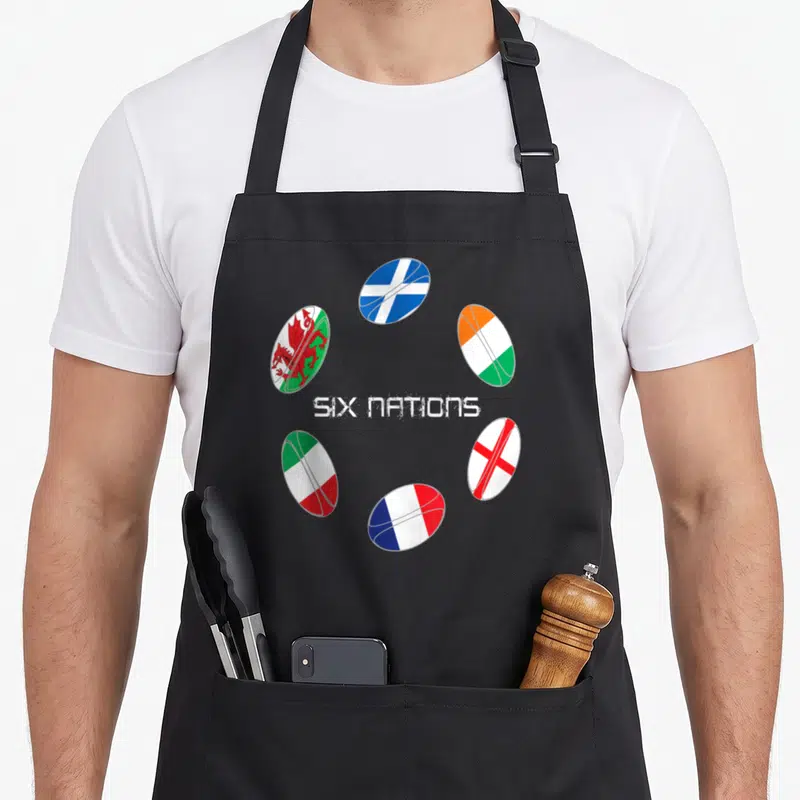 Tablier de cuisine noir avec six ballons de rugby aux couleurs des drapeaux nationaux disposés en cercle autour d'un texte blanc.
