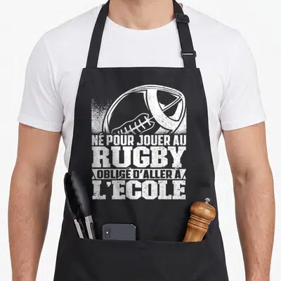 Tablier de cuisine noir avec illustration de ballon de rugby et texte humoristique sur l'école en typographie blanche usée.