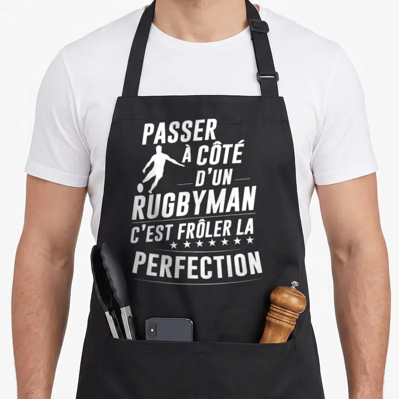 Tablier de cuisine noir avec une silhouette blanche de joueur de rugby et le texte blanc passant à côté d'un rugbyman c'est frôler la perfection.