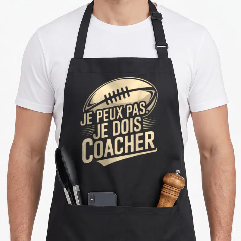 Tablier de cuisine noir avec illustration de ballon de rugby et texte Je peux pas je dois coacher en beige vintage.