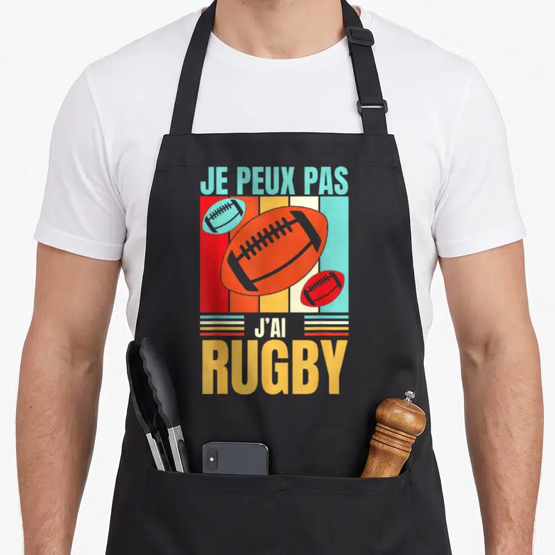 Tablier de cuisine noir avec motif rétro affichant des ballons de rugby orange et le texte Je peux pas j'ai rugby en lettres colorées.
