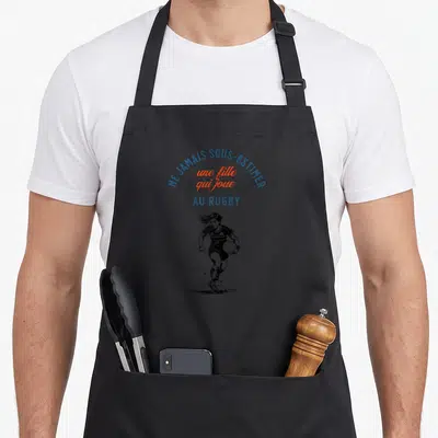 Tablier de cuisine noir avec une illustration d'une joueuse de rugby courant et le texte ne jamais sous-estimer une fille qui joue au rugby.