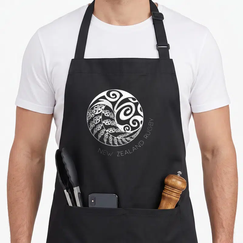 Tablier de cuisine noir avec un logo circulaire blanc aux motifs maoris et l'inscription New Zealand Rugby en bas.
