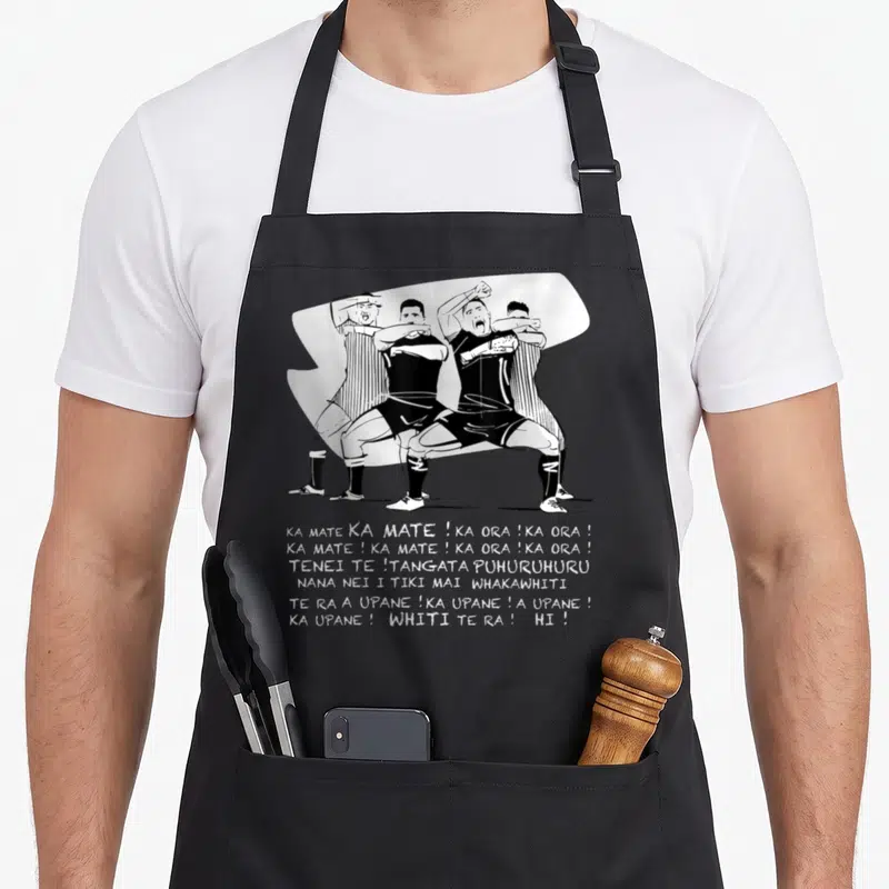 Tablier de cuisine noir avec illustration en noir et blanc de joueurs effectuant un haka et des paroles imprimées en dessous.