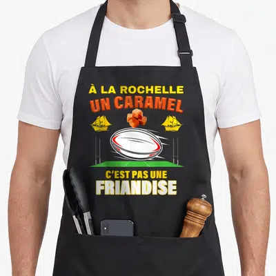 Tablier de cuisine noir avec un ballon de rugby, des caramels et deux navires jaunes sur fond vert.