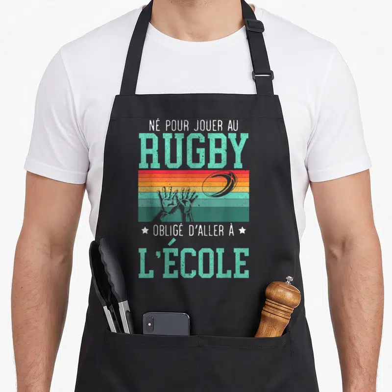 Tablier de cuisine noir avec inscription né pour jouer au rugby obligé d'aller à l'école sur fond de bandes colorées.