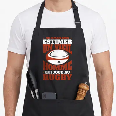 Tablier de cuisine noir avec texte Ne jamais sous-estimer un vieil homme qui joue au rugby et un ballon de rugby central.