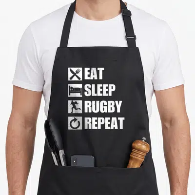 Tablier de cuisine noir avec inscription blanche Eat Sleep Rugby Repeat et icônes stylisées verticales.
