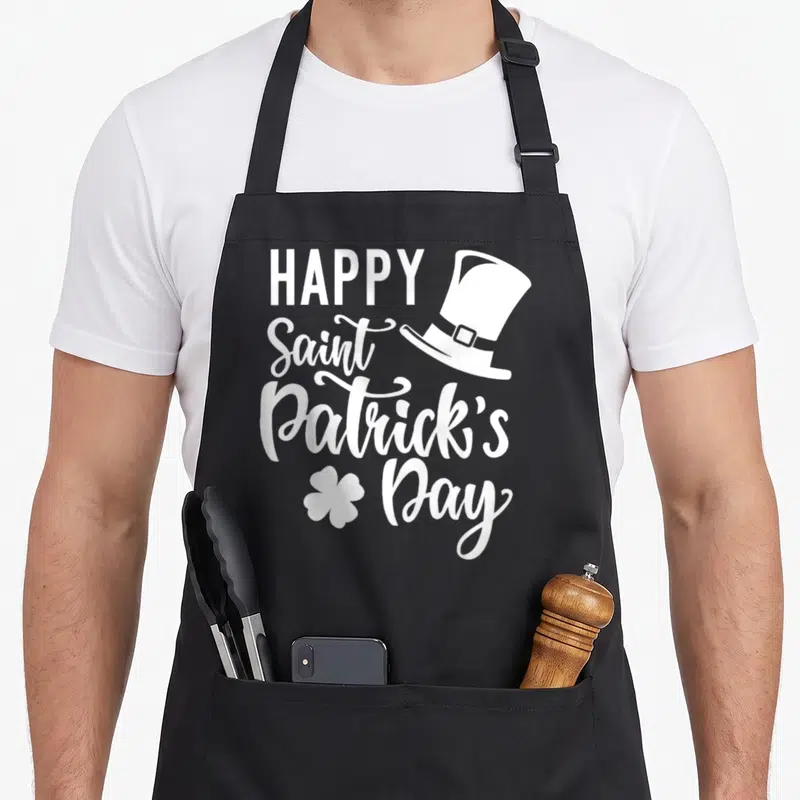 Tablier de cuisine noir avec inscription Happy Saint Patrick's Day, chapeau haut de forme et trèfle à quatre feuilles en blanc.