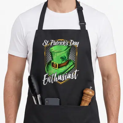 Tablier de cuisine noir avec illustration chapeau vert trèfle et texte St. Patrick's Day Enthusiast encadré d'or.