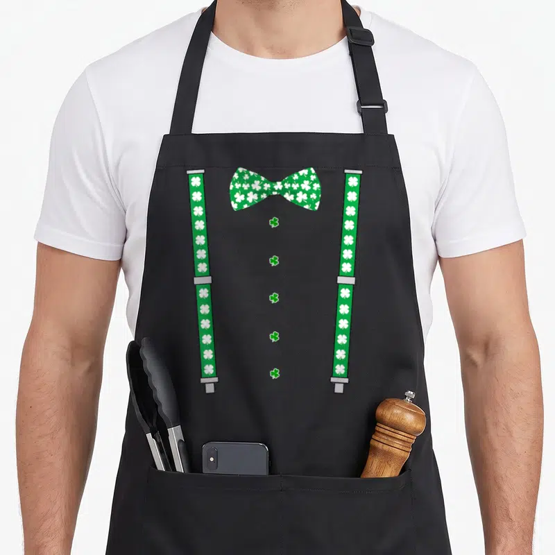 Tablier de cuisine noir avec illustration de bretelles vertes et noeud papillon à motifs de trèfles blancs.