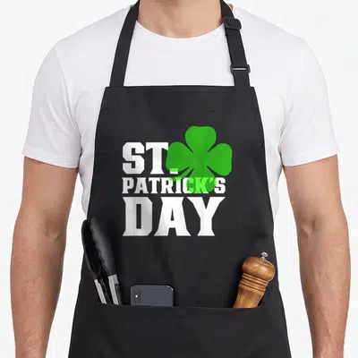 Tablier de cuisine noir avec inscription blanche St Patrick's Day et motif de trèfle vert à quatre feuilles sur le devant.