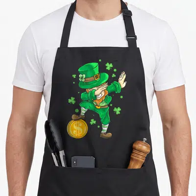 Tablier de cuisine noir avec illustration colorée d'un lutin irlandais effectuant un mouvement de danse dab au-dessus d'une pièce dorée.