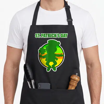 Tablier de cuisine noir avec une silhouette de lutin vert tenant un trèfle sur fond de bandes colorées.