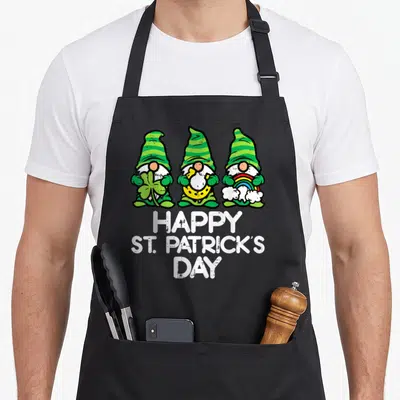 Tablier de cuisine noir avec trois gnomes verts tenant un trèfle, une pièce d'or et un arc-en-ciel sous le texte Happy St Patrick's Day.